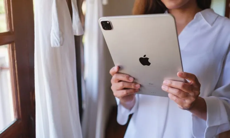 Woman holds a silver iPad near a window with white curtains.