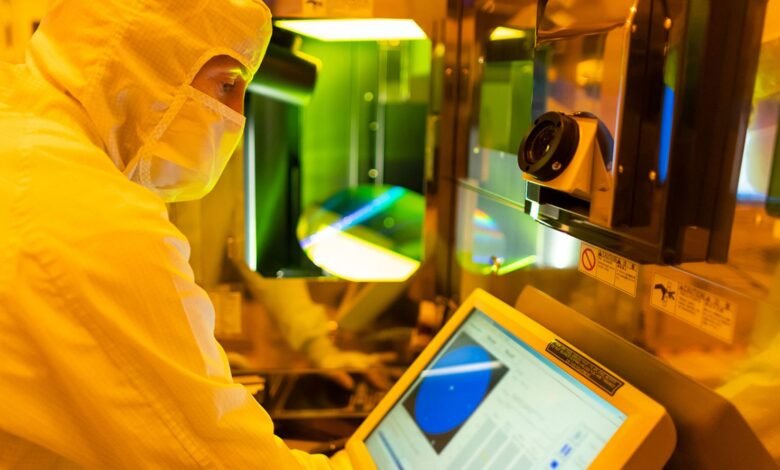 Technician in cleanroom suit inspecting a silicon wafer in a manufacturing environment.