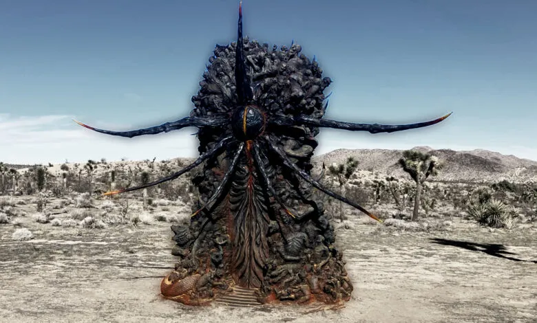 Dark, intricate statue with figures in desert landscape under a blue sky.