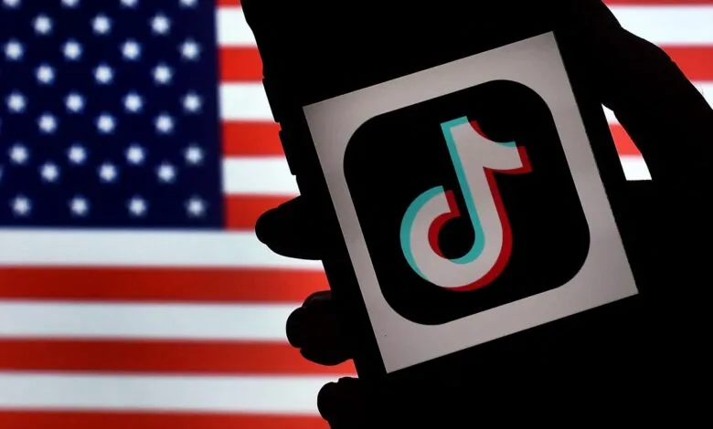Hand holding phone with TikTok logo against blurred US flag backdrop.