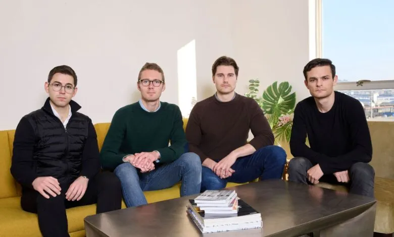 Four young men sit on a yellow couch in a bright, modern office space.