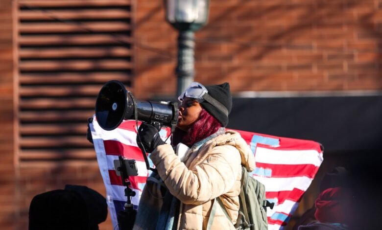 Person speaks into megaphone at outdoor protest, American flag visible.