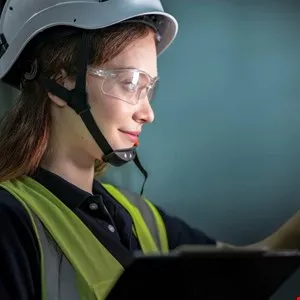 Woman in hard hat and safety vest holding clipboard, looking to the side.