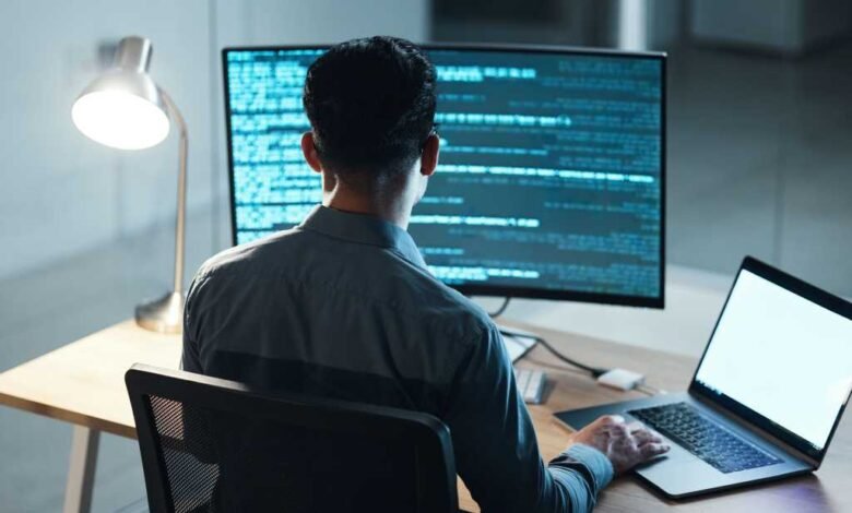 Person coding at desk with laptop and monitor displaying code.