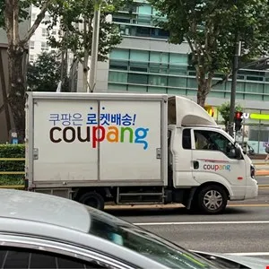 Coupang delivery truck on a city street with modern buildings in the background.