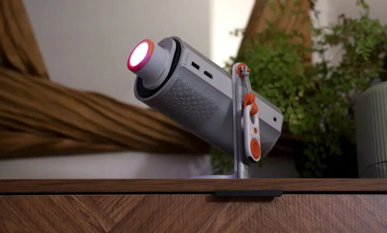 Portable projector on stand with red light, plants in background.