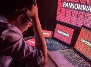 Person with head in hands surrounded by screens displaying 'RANSOMWARE' in red.