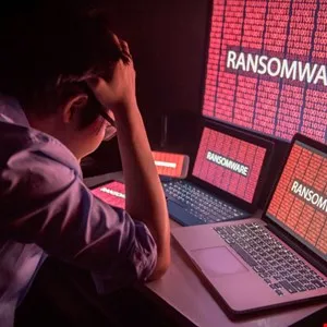 Person with head in hands surrounded by screens displaying 'RANSOMWARE' in red.