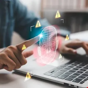 Person interacts with a laptop displaying a digital face and warning symbols.