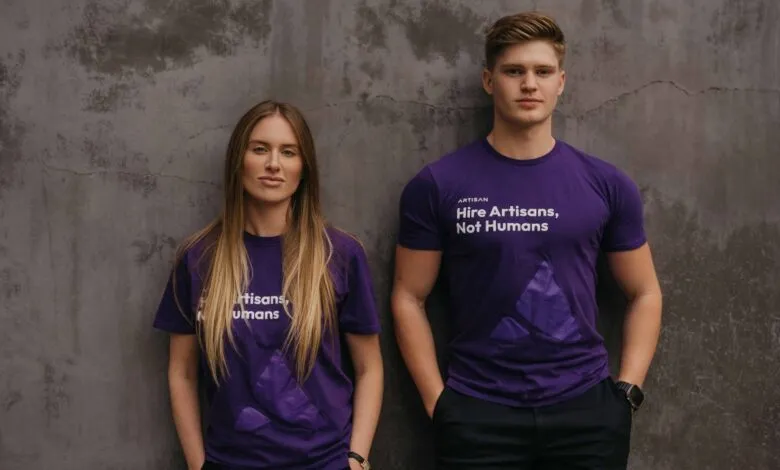 Two people wearing purple Artisan t-shirts stand against a textured gray wall.