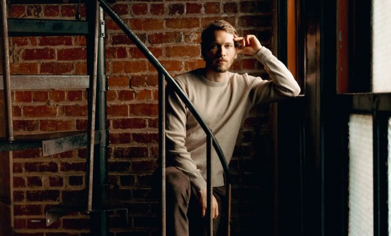 Man with beard poses on spiral staircase against brick wall.