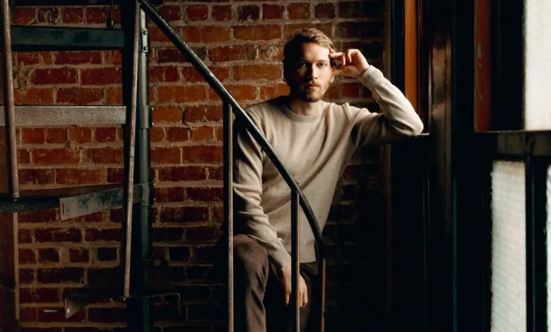 Man with beard poses on spiral staircase against brick wall.