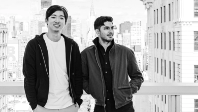 Two men stand against a NYC skyline in a black and white portrait.