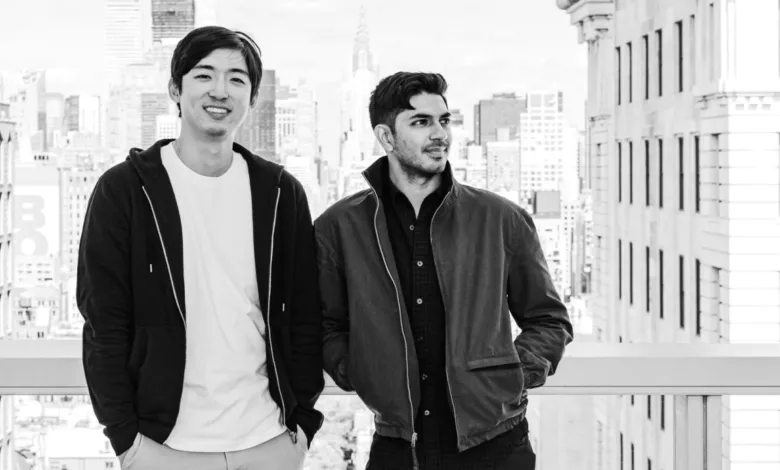 Two men stand against a NYC skyline in a black and white portrait.