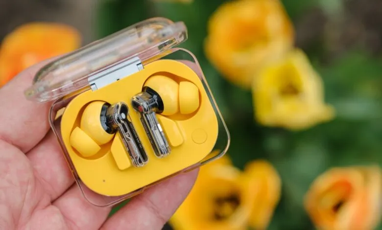 Hand holding yellow Nothing Ear (stick) earbuds in clear charging case.