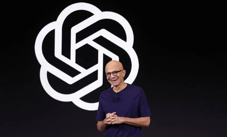Satya Nadella smiles on stage with OpenAI logo in background.