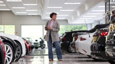 Woman in blazer stands in car showroom holding phone, looking at vehicles.