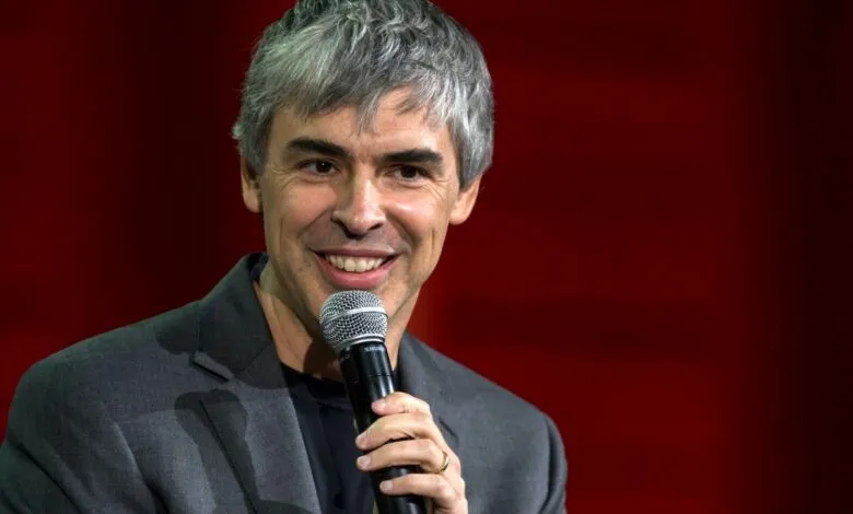 Larry Page smiles while holding a microphone against a red backdrop.