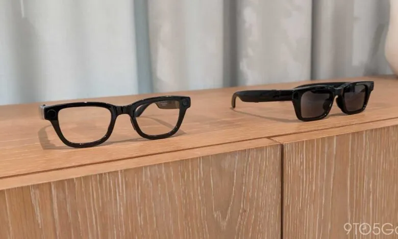 Two pairs of black smart glasses on a light wood surface.