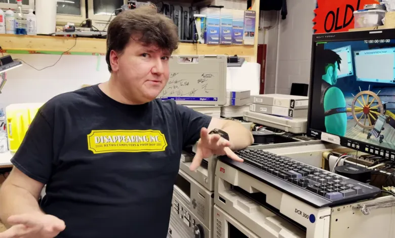 Man in 'Disappearing Inc.' shirt stands near vintage computer equipment.