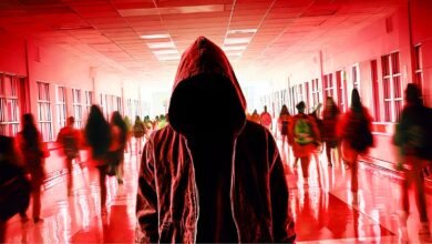 Figure in hoodie stands in school hallway with blurred students.