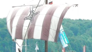Replica Viking ship with striped sail and emblem sailing on water.