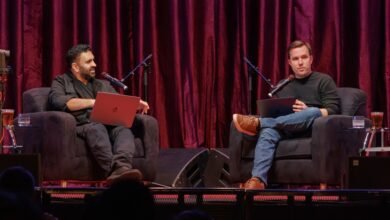 Two men sit in armchairs on stage, holding laptops and speaking into microphones.