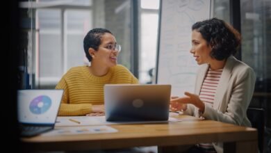 Two women collaborate at a desk with laptops and charts in a modern office.