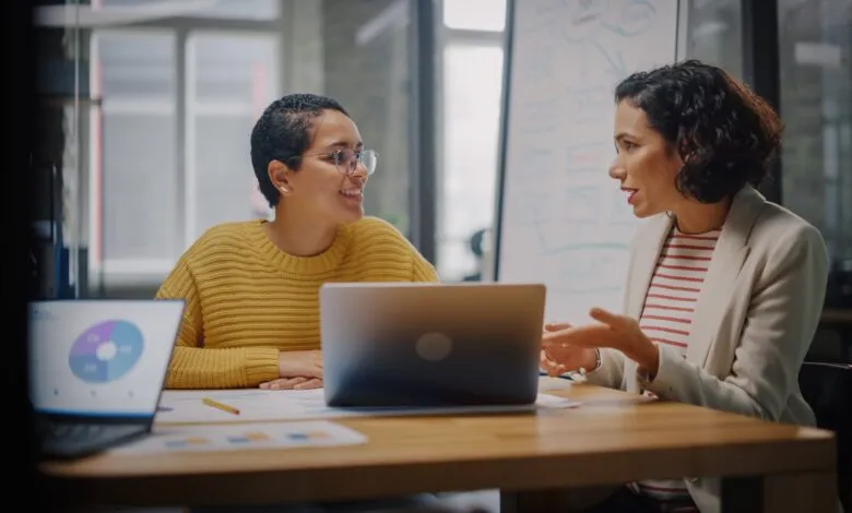 Two women collaborate at a desk with laptops and charts in a modern office.