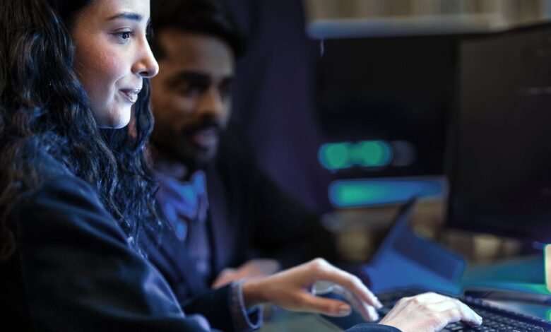 Woman coding at computer with colleague in dark office setting.