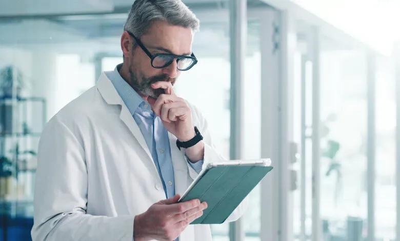 Doctor in lab coat thoughtfully reviews tablet data in bright office.