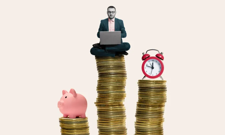 Man with laptop sits atop coins, next to piggy bank and alarm clock.