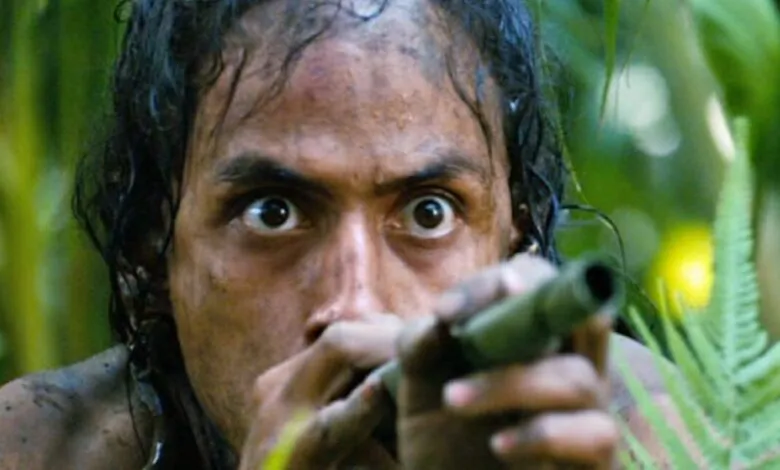 Close-up of a man aiming a blowgun in a jungle setting.