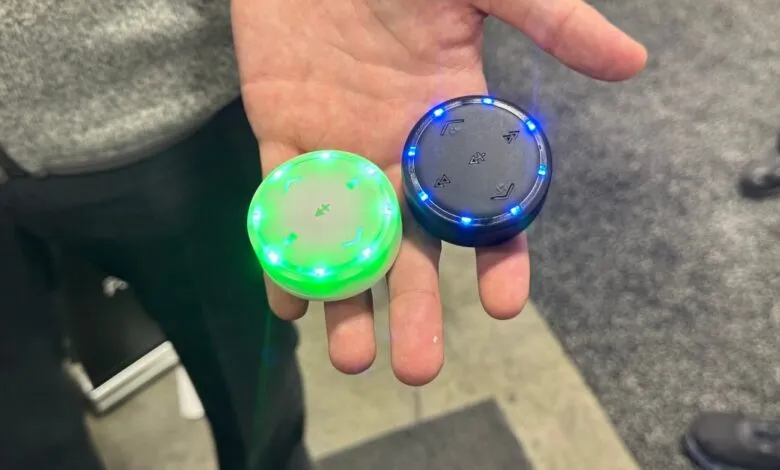 Hand holding two round Auracast transceivers, one green, one blue.