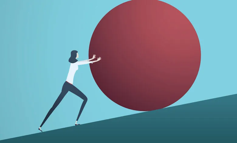 Businesswoman pushes a large red boulder uphill, symbolizing a difficult challenge.