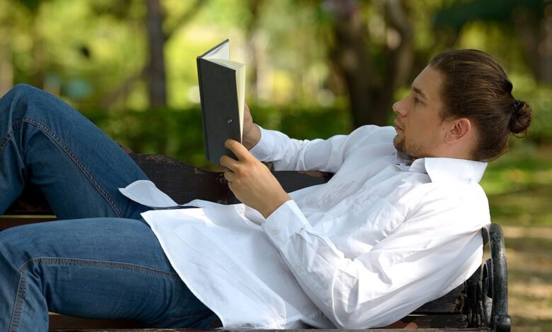 Man reading book on park bench, wearing jeans and white shirt.