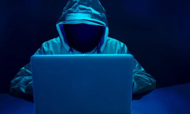 Person in hoodie sits at laptop in dark, blue-lit room.
