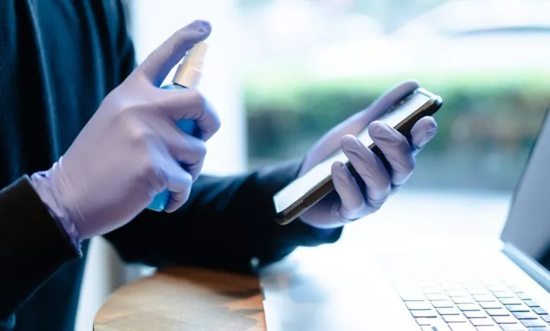 Person in gloves sanitizing phone near laptop.
