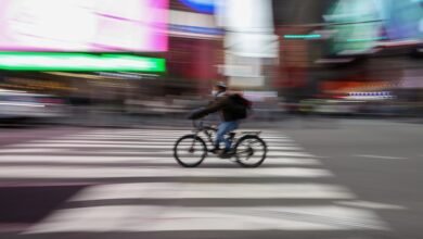 Cyclist wearing mask rides bike across crosswalk in blurred city scene.