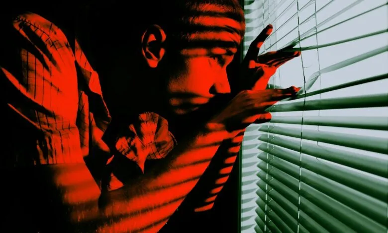 Man peers through blinds, bathed in red light, creating a sense of mystery.