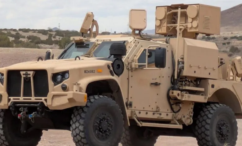 Tan military vehicle with advanced sensor equipment on a desert landscape.