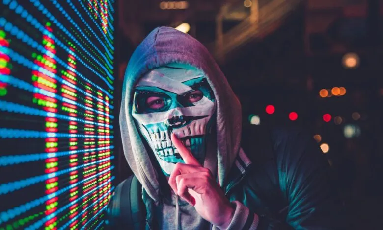 Person in skull mask and hoodie stands before a screen of data.