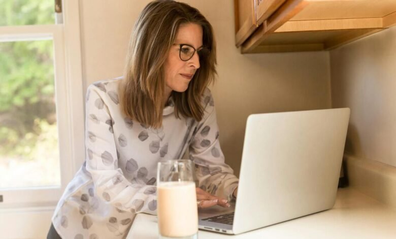 Woman with glasses working on laptop in a bright kitchen.