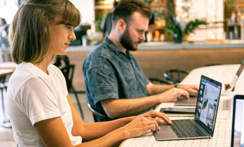 Two people working on laptops at a table in a bright, modern cafe.