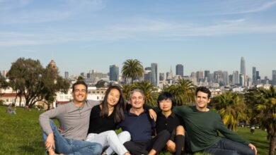 Five smiling people sit on a grassy hill with San Francisco skyline in background.