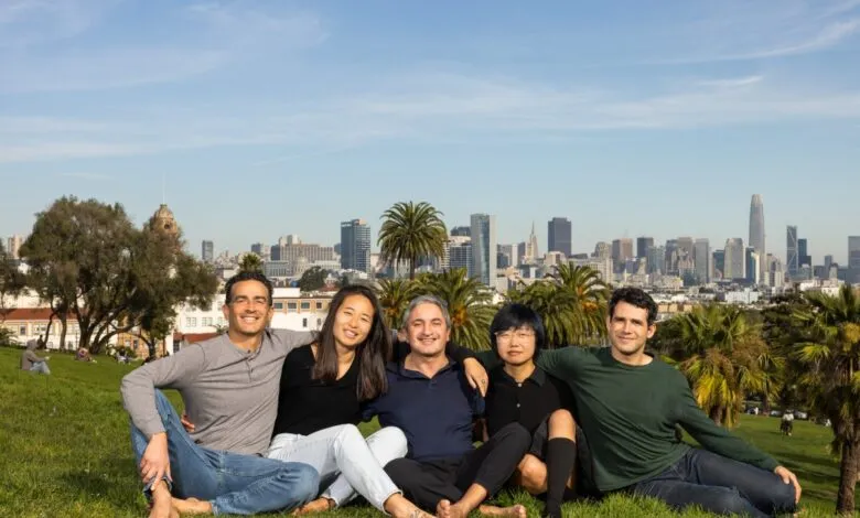 Five smiling people sit on a grassy hill with San Francisco skyline in background.