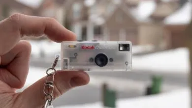 Hand holds a Kodak Charmera camera keychain against a blurred outdoor background.
