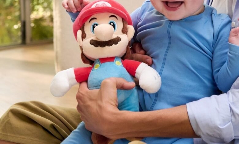 Baby in blue outfit holds a Mario plush toy, sitting on someone's lap.