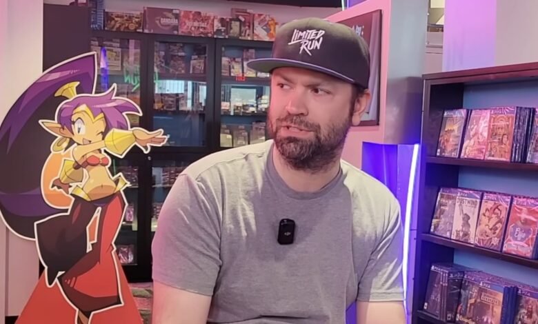 Man in Limited Run hat sits next to Shantae standee in game store.