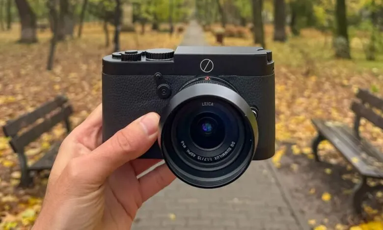 Hand holds a black Leica camera in a park setting with benches and autumn leaves.